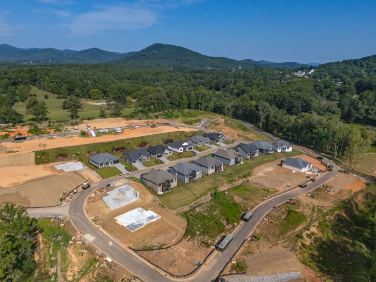 Aerial view of a residential development nestled in a lush, forested landscape. The image shows a community of newly constructed homes with dark roofs surrounded by rolling hills, mature trees, and winding roads. Construction equipment and cleared land indicate an ongoing development project. The scenic mountain backdrop adds to the picturesque setting, creating an appealing residential environment.