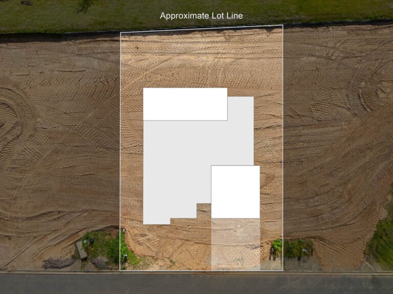 An aerial view of a construction site, with an approximate lot line marked by a white rectangular outline. The ground is covered in textured soil and gravel, with some vegetation visible around the edges. The image provides a clear visual reference for the planned layout and dimensions of the construction project.