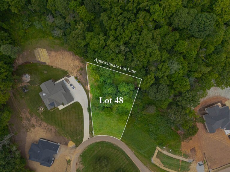 Aerial view of a residential lot surrounded by lush green forest. The lot, labeled as Lot 48, is outlined with an "Approximate Lot Line" marker. The image showcases the property's natural setting, with the forested area contrasting with the cleared land and nearby homes.