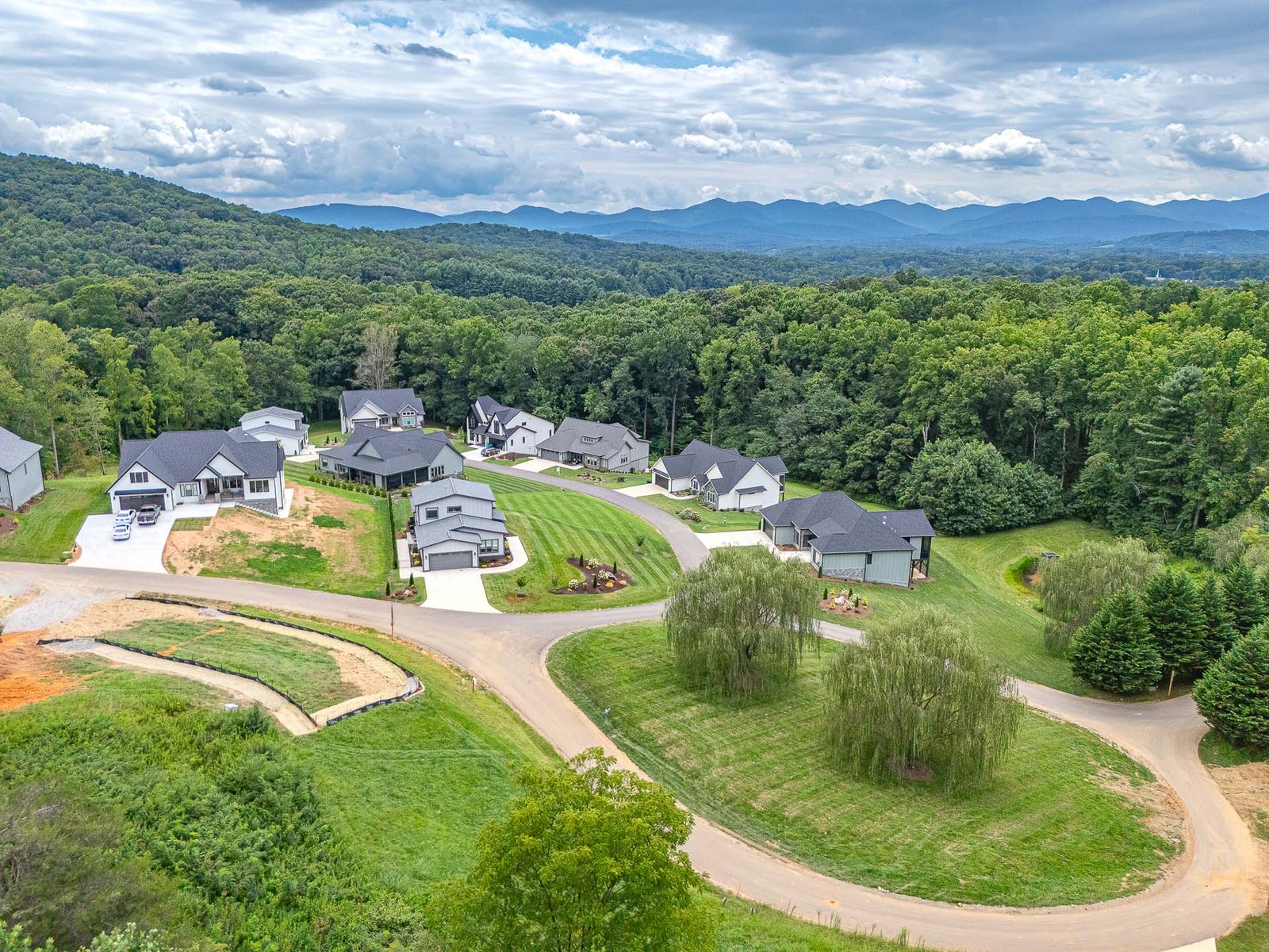 An aerial view of a residential neighborhood nestled in a lush, forested landscape. The image shows a community of modern, well-maintained homes with neatly manicured lawns, surrounded by a backdrop of rolling hills and mountains in the distance. The winding roads and pathways weave through the verdant greenery, creating a picturesque and serene setting.