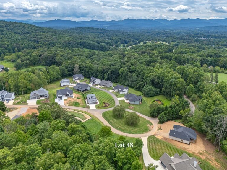 Aerial view of a residential community nestled in a lush, forested landscape. The image shows a development with multiple homes surrounded by rolling hills, dense greenery, and winding roads. The mountainous backdrop adds to the scenic, serene atmosphere of the setting.