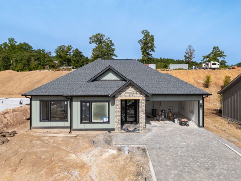 A newly constructed modern single-story house with a gray shingled roof, stone exterior, and large windows. The house is surrounded by a dirt construction site with trees in the background, suggesting it is a new home in a residential development.