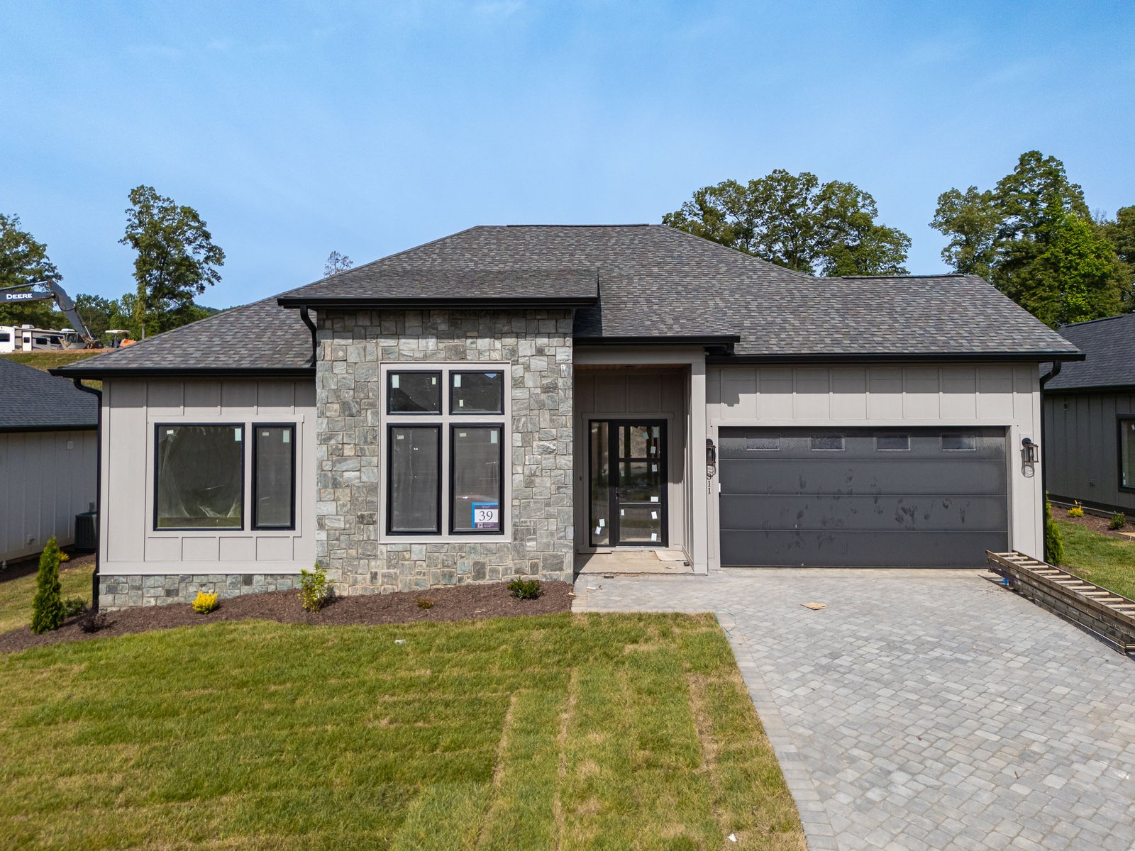 A modern, two-story house with a gray exterior and a stone facade. The home features large windows, a garage door, and a paved driveway leading to the entrance. Surrounded by lush greenery and trees, the house exudes a warm and inviting atmosphere.