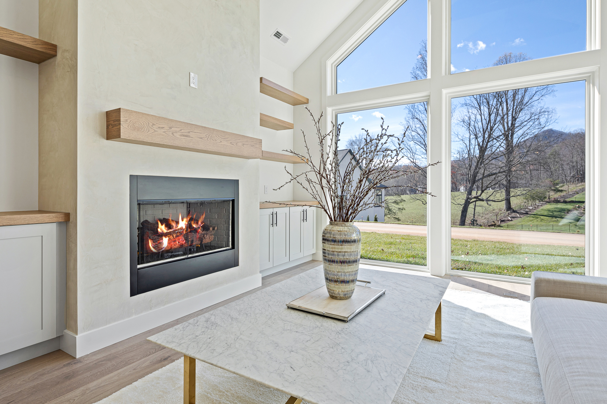 A spacious and modern living room with a large window overlooking a scenic outdoor landscape. The room features a cozy fireplace surrounded by wooden beams, and a stylish vase with branches on a marble coffee table, creating a warm and inviting atmosphere.