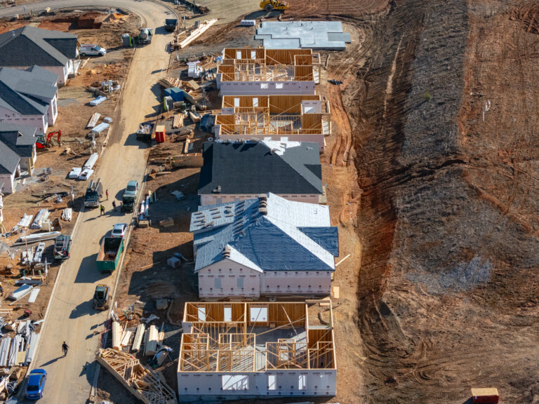 Aerial view of an active construction site with partially completed buildings, construction materials, and heavy machinery scattered throughout the dirt and gravel landscape. The scene depicts the early stages of a large-scale residential or commercial development project underway.