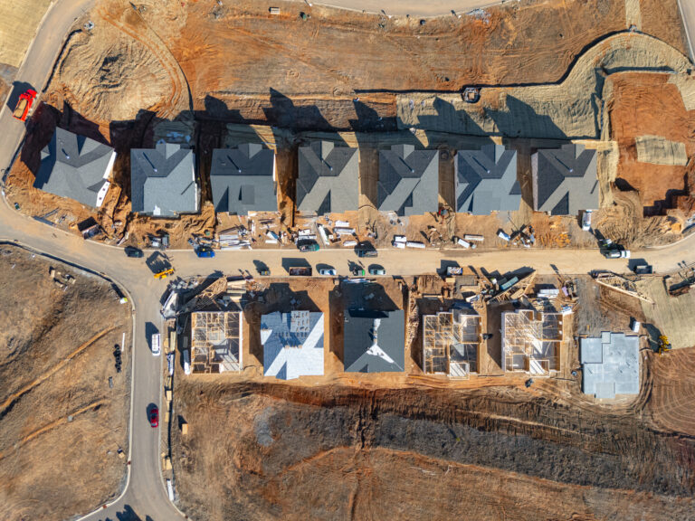 Aerial view of a construction site with various buildings and structures in various stages of development. The image shows a large open area with earthmoving equipment and materials scattered throughout, as well as several partially constructed buildings with distinctive geometric patterns on their facades.