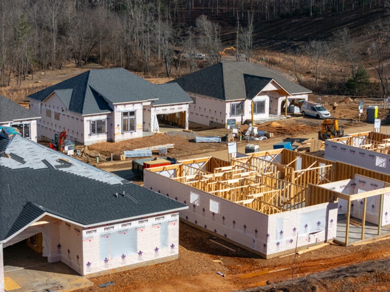 A construction site with partially completed residential buildings, including framed structures, roofing, and exterior siding. The scene depicts an active building project in a wooded, rural setting, with construction equipment and materials visible on the site.