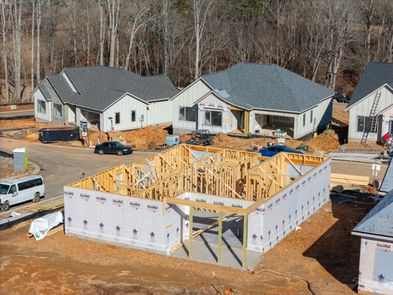A construction site featuring partially built houses with wooden frames and gray roofs, surrounded by a wooded area. The site is filled with construction materials, equipment, and vehicles, indicating an ongoing residential development project.