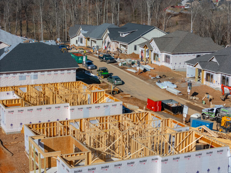 A bustling construction site with partially built wooden frames of new homes, surrounded by heavy machinery, construction materials, and workers actively engaged in the building process. The scene depicts the early stages of a residential development project, with the framing and foundation work underway amidst a wooded, hilly landscape.