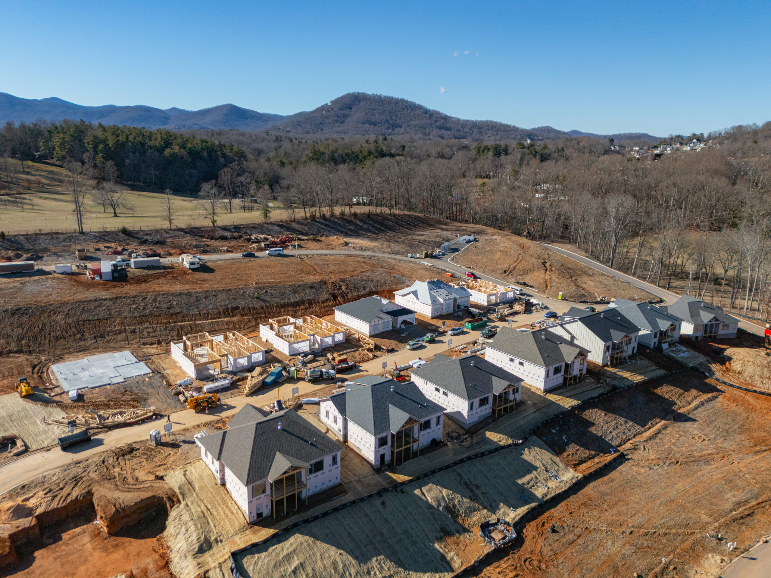 Aerial view of a residential construction site nestled in a scenic mountain landscape. The image shows partially completed homes, construction equipment, and cleared land surrounded by forested hills and valleys. The scene depicts an active development project in a picturesque rural setting.
