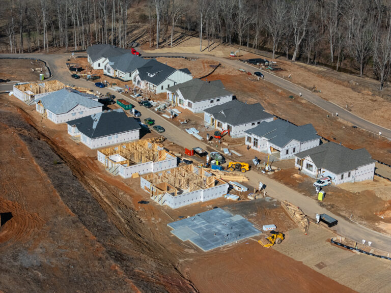 Aerial view of a residential construction site with multiple partially built homes, construction equipment, and workers actively engaged in the building process. The scene depicts an active housing development in a wooded, rural area with a dirt road running through the center.