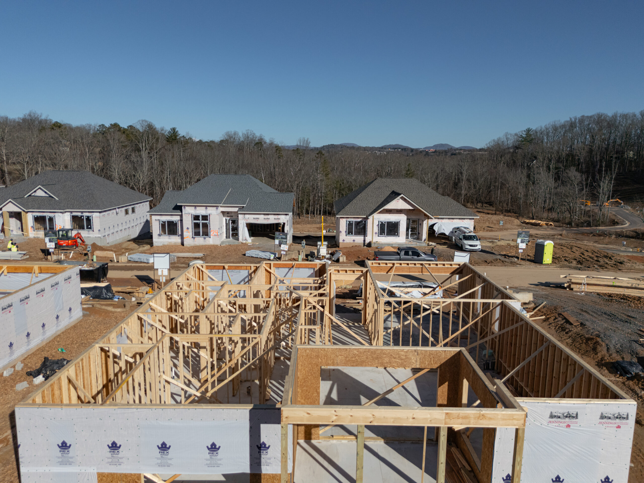 A construction site with multiple new homes under construction. The image shows the framing and foundations of several houses, surrounded by building materials and equipment. In the background, there is a forested area with hills visible, indicating a rural or suburban setting. The scene depicts the early stages of a residential development project.