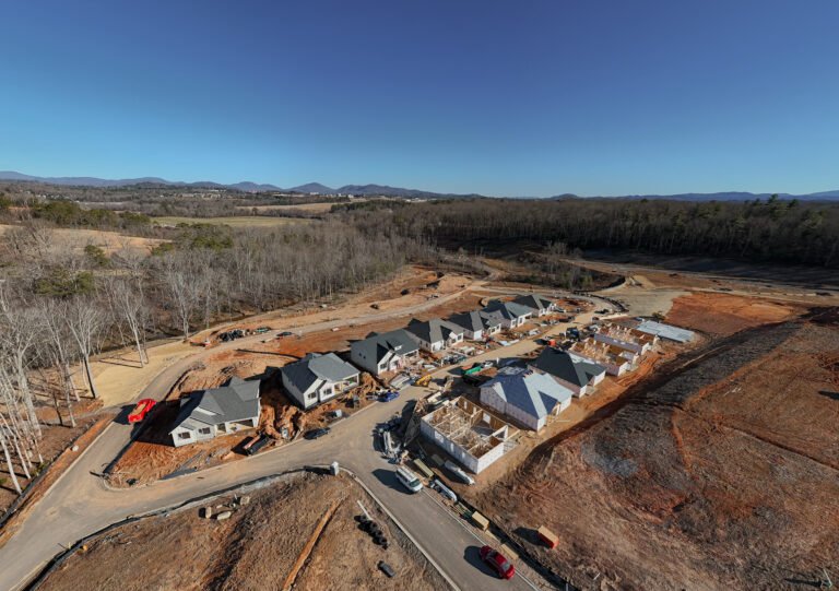 An aerial view of a residential construction site nestled in a rural landscape, with partially built homes, construction equipment, and a winding road leading through the development. The surrounding area features lush forests, rolling hills, and a clear blue sky, creating a picturesque setting for the new community.