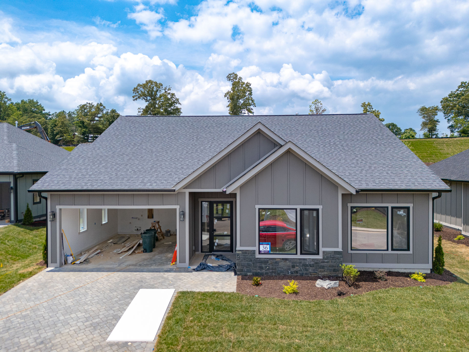 A modern, single-story house with a gray exterior, a gabled roof, and large windows. The house is surrounded by lush greenery, including trees and well-manicured landscaping. The driveway is paved with brick pavers, and the home appears to be in the final stages of construction or renovation.