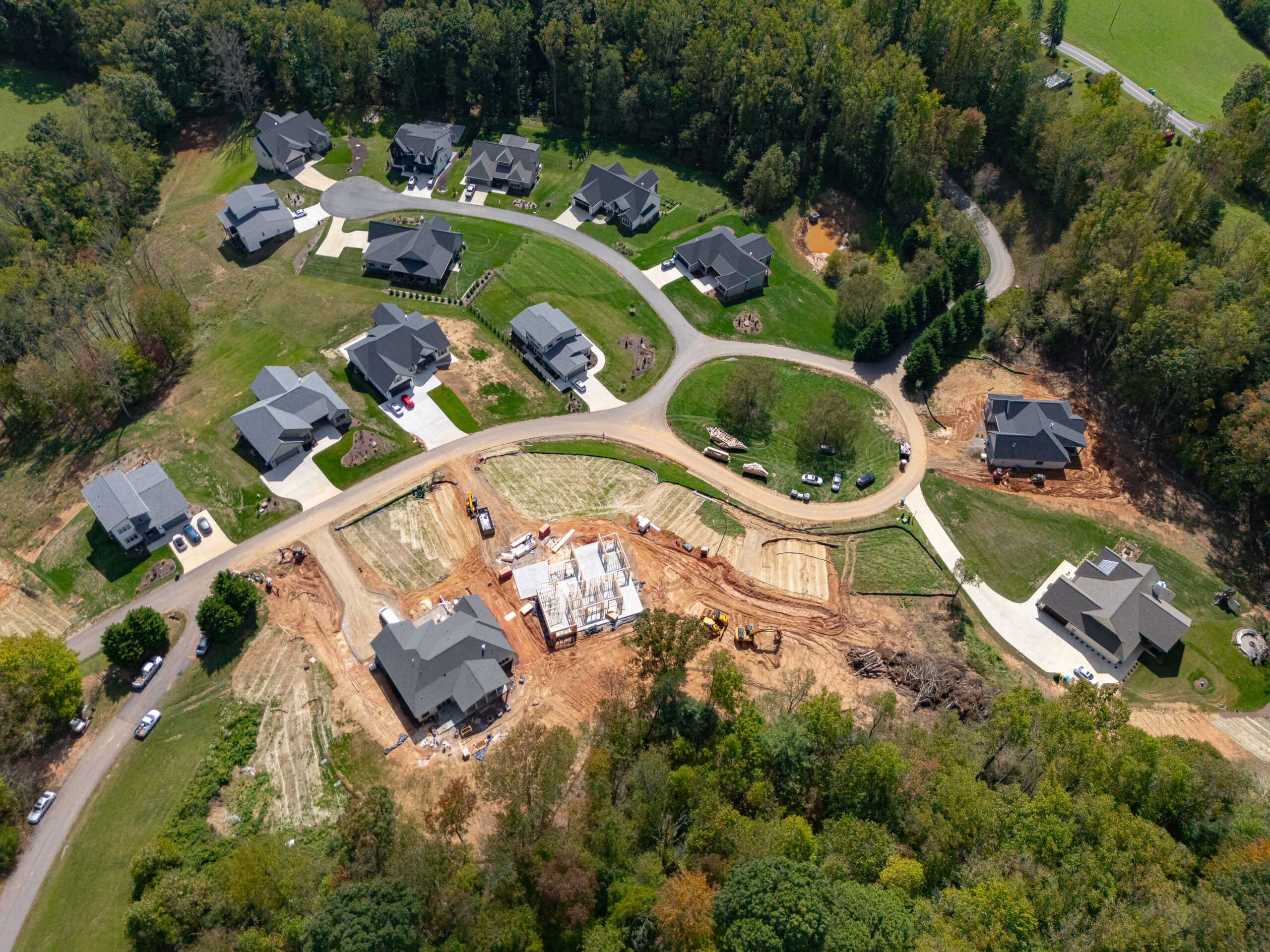 Aerial view of a residential neighborhood under construction, with newly built homes surrounded by lush greenery and winding roads. The image shows a mix of completed and partially constructed houses, with construction equipment and materials visible on the site. The community appears to be situated in a picturesque, wooded area, creating a serene and natural setting for the new development.