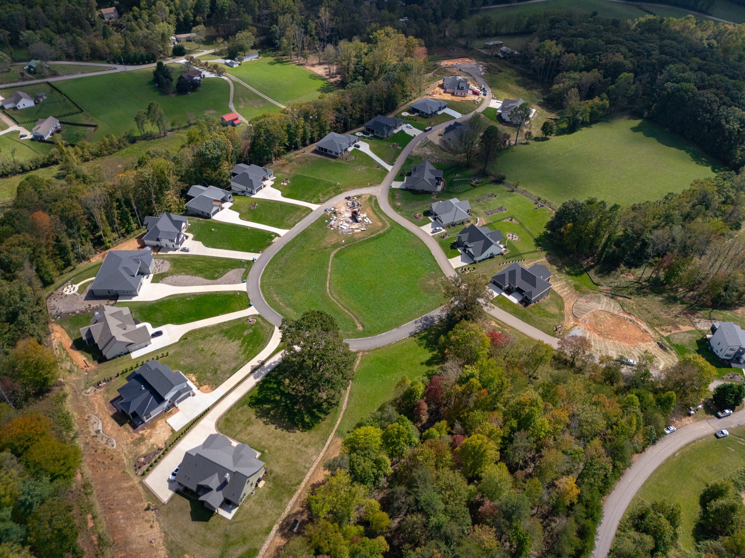 Aerial view of a residential neighborhood nestled in a lush, forested landscape. The community features a network of winding roads, large homes with dark roofs, and well-manicured green spaces. The scene showcases the harmonious integration of modern architecture and natural surroundings, creating a picturesque and serene suburban setting.