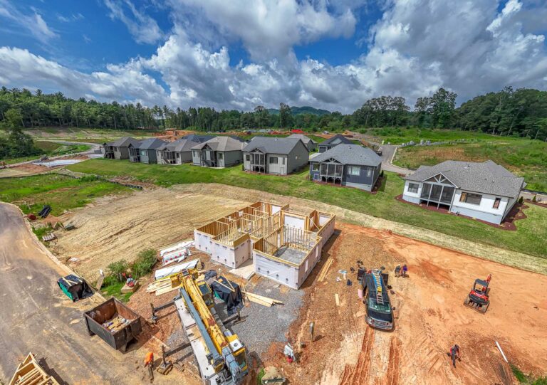 A sprawling residential development under construction, with partially built homes, heavy machinery, and construction materials scattered throughout the site. The scene is surrounded by a lush, forested landscape with a cloudy blue sky overhead, creating a picturesque backdrop for the ongoing development.