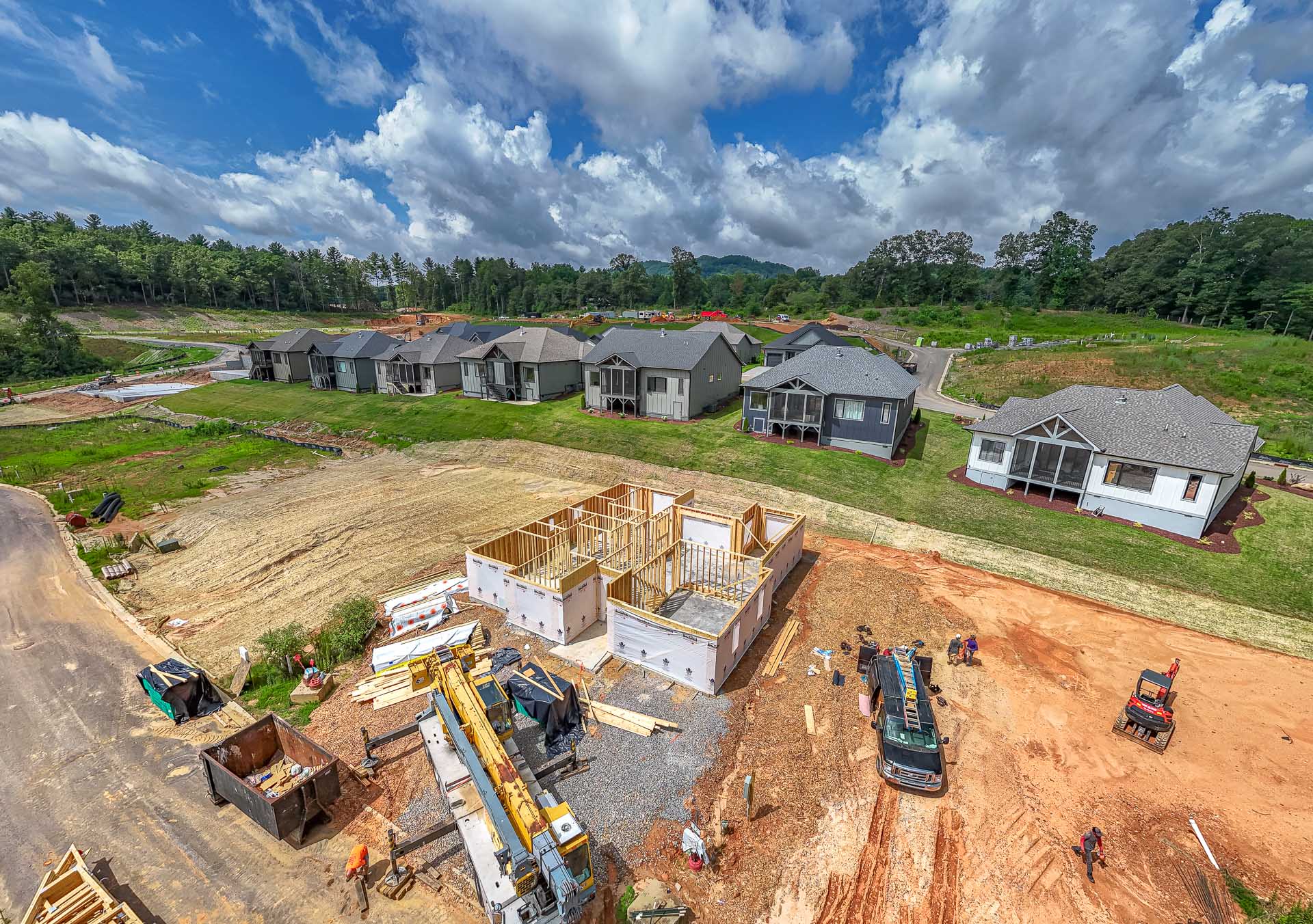 A sprawling residential development under construction, with partially built homes, heavy machinery, and construction materials scattered throughout the site. The scene is surrounded by a lush, forested landscape with a cloudy blue sky overhead, creating a picturesque backdrop for the ongoing development.