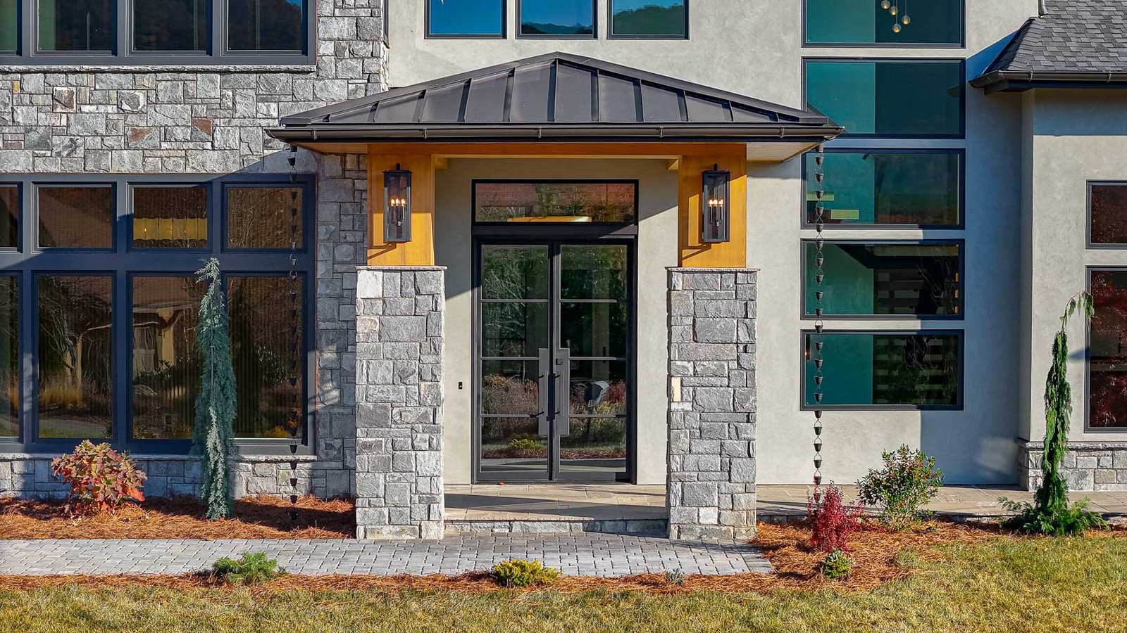 A modern, two-story residential building with a stone and stucco exterior, featuring a prominent entryway with a gabled roof and decorative lighting fixtures. The building is surrounded by lush landscaping, including a paved walkway and various plants and shrubs. The large windows provide ample natural light and offer scenic views of the surrounding environment.