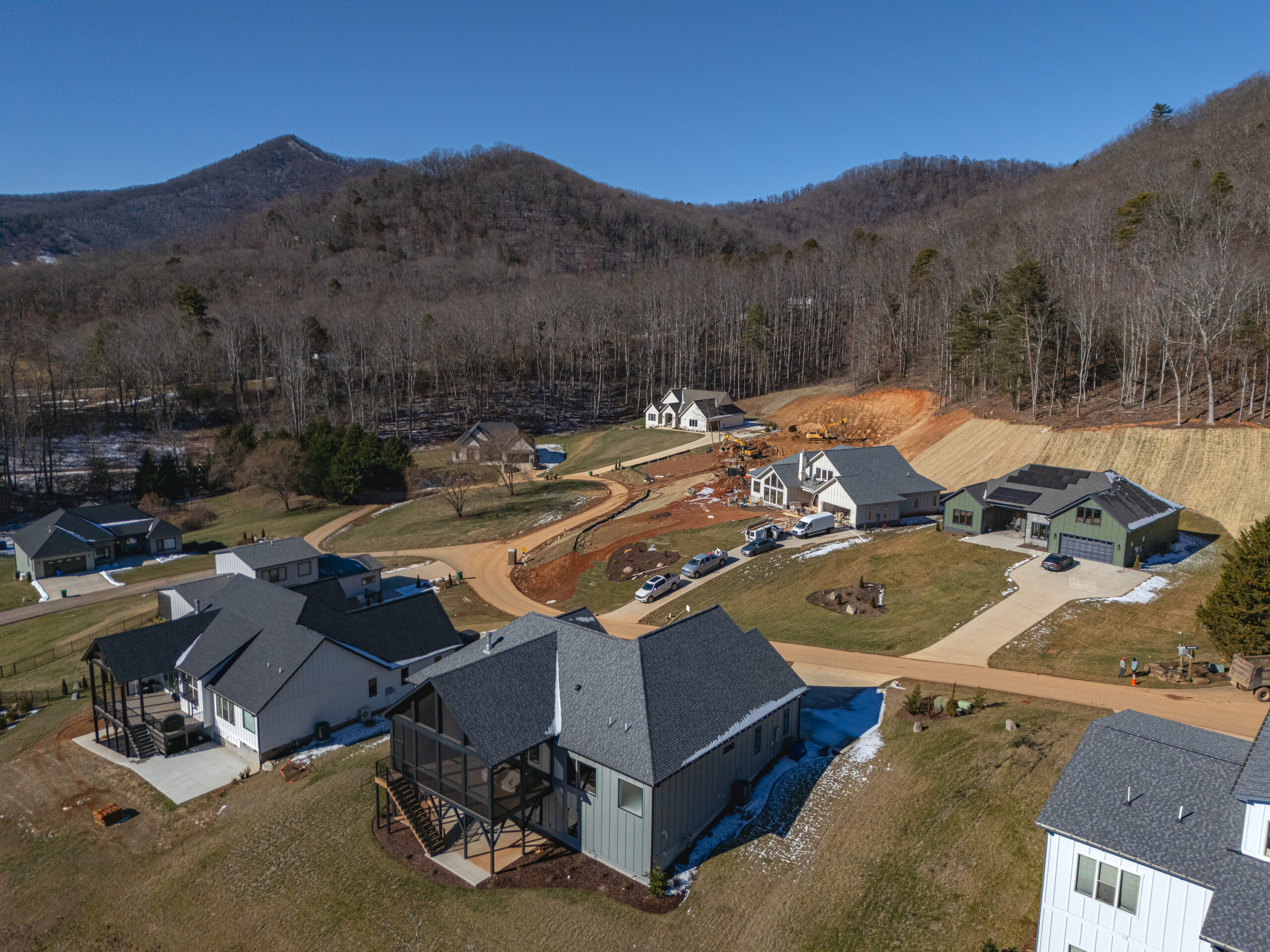 An aerial view of a residential neighborhood nestled in a mountainous, forested landscape. The image shows a cluster of modern, well-appointed homes with slate roofs and attached garages, surrounded by lush greenery and a winding dirt road. In the background, the rolling hills and dense tree cover create a picturesque, rural setting.