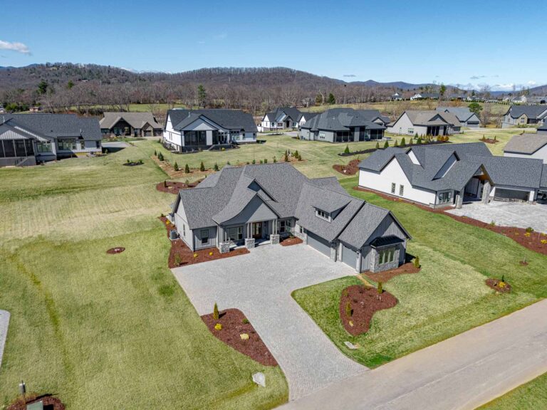 A sprawling residential community nestled in a lush, verdant landscape, featuring a collection of well-designed, modern homes with gray roofs and stone accents, surrounded by meticulously landscaped yards, winding pathways, and a backdrop of rolling hills and a clear blue sky.