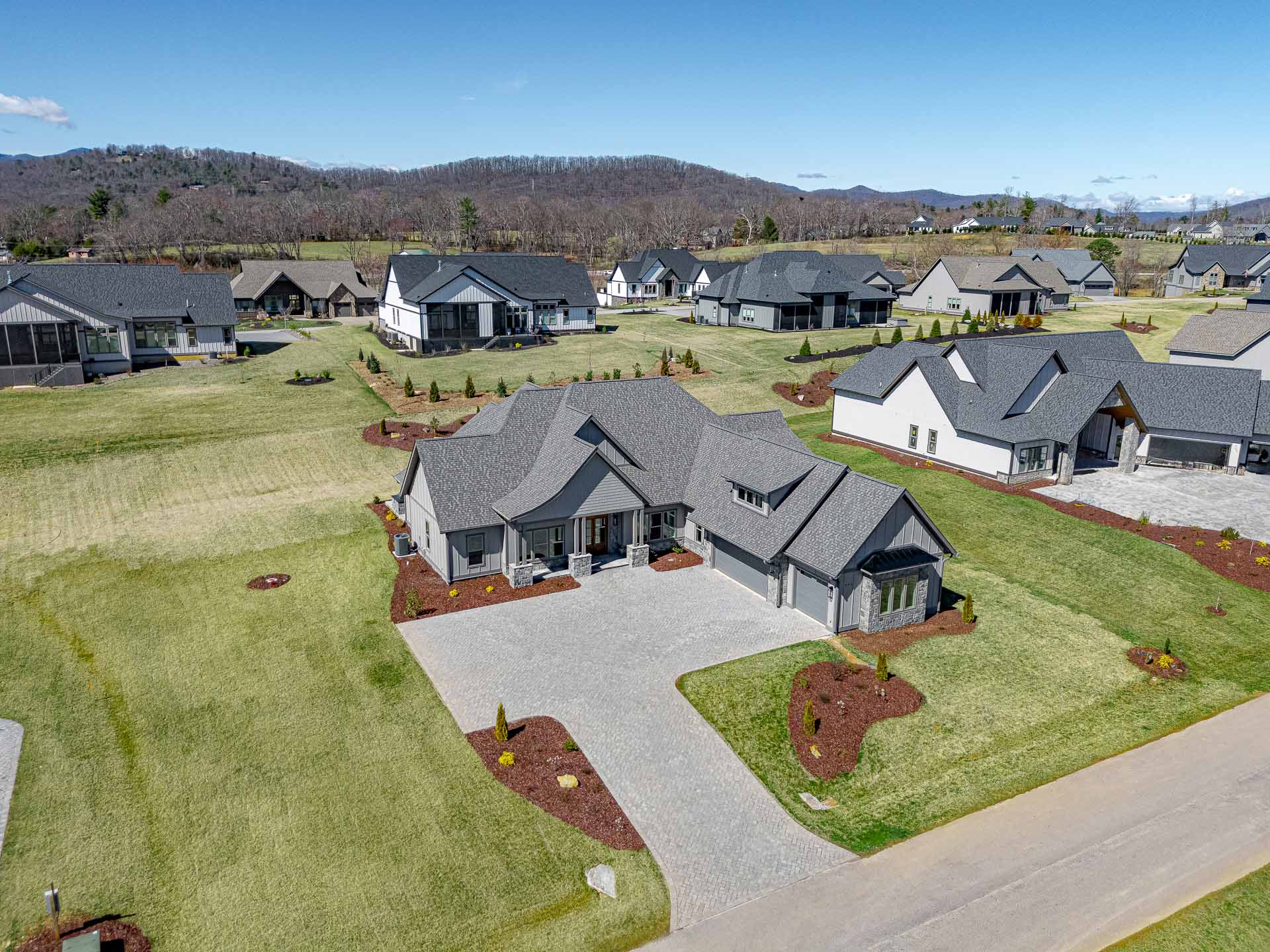 A sprawling residential community nestled in a lush, verdant landscape, featuring a collection of well-designed, modern homes with gray roofs and stone accents, surrounded by meticulously landscaped yards, winding pathways, and a backdrop of rolling hills and a clear blue sky.