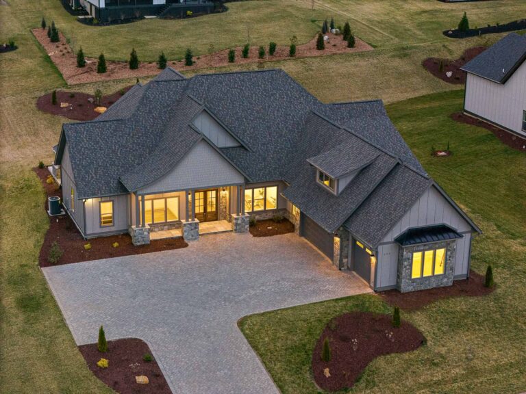 Aerial view of a large, modern two-story home with a gray slate roof, stone accents, and a wrap-around porch. The home is surrounded by a well-manicured lawn with landscaped flower beds and evergreen trees, creating a picturesque and inviting outdoor setting.