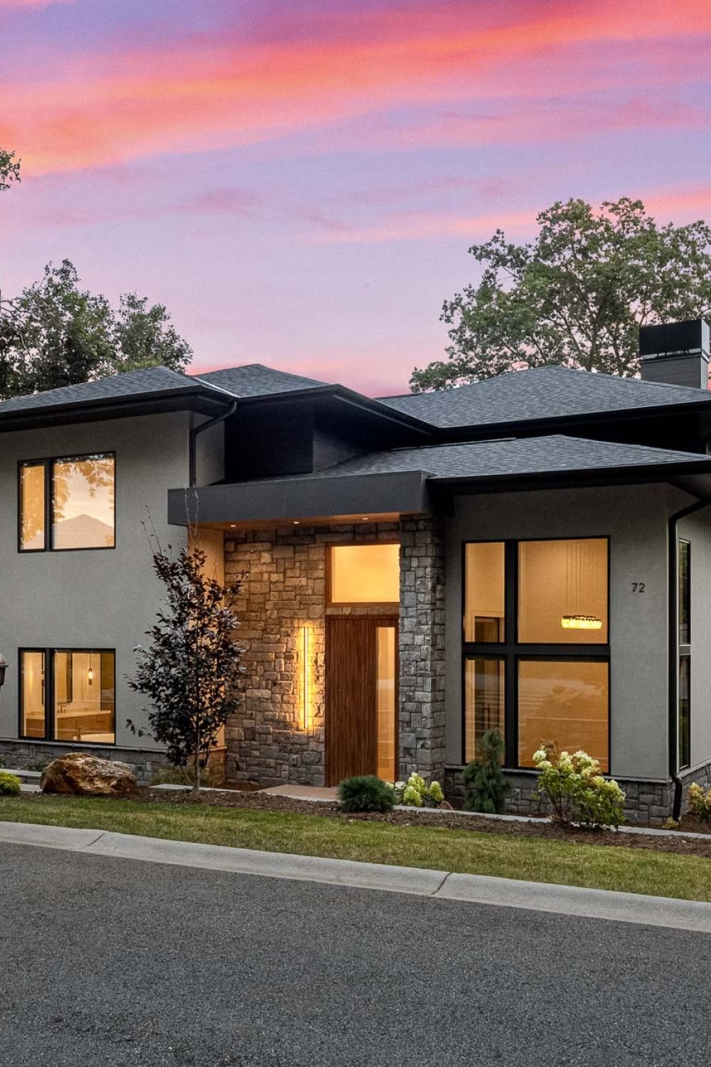 A modern two-story house with a stone exterior, large windows, and a wooden front door. The house is surrounded by lush greenery and set against a vibrant sunset sky, creating a warm and inviting atmosphere. The landscaping features well-manicured shrubs and trees, adding to the property's curb appeal.